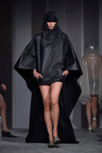 Model walking a fashion show in a black hooded cape with wide sleeves and short inner garment visible at the hembed.