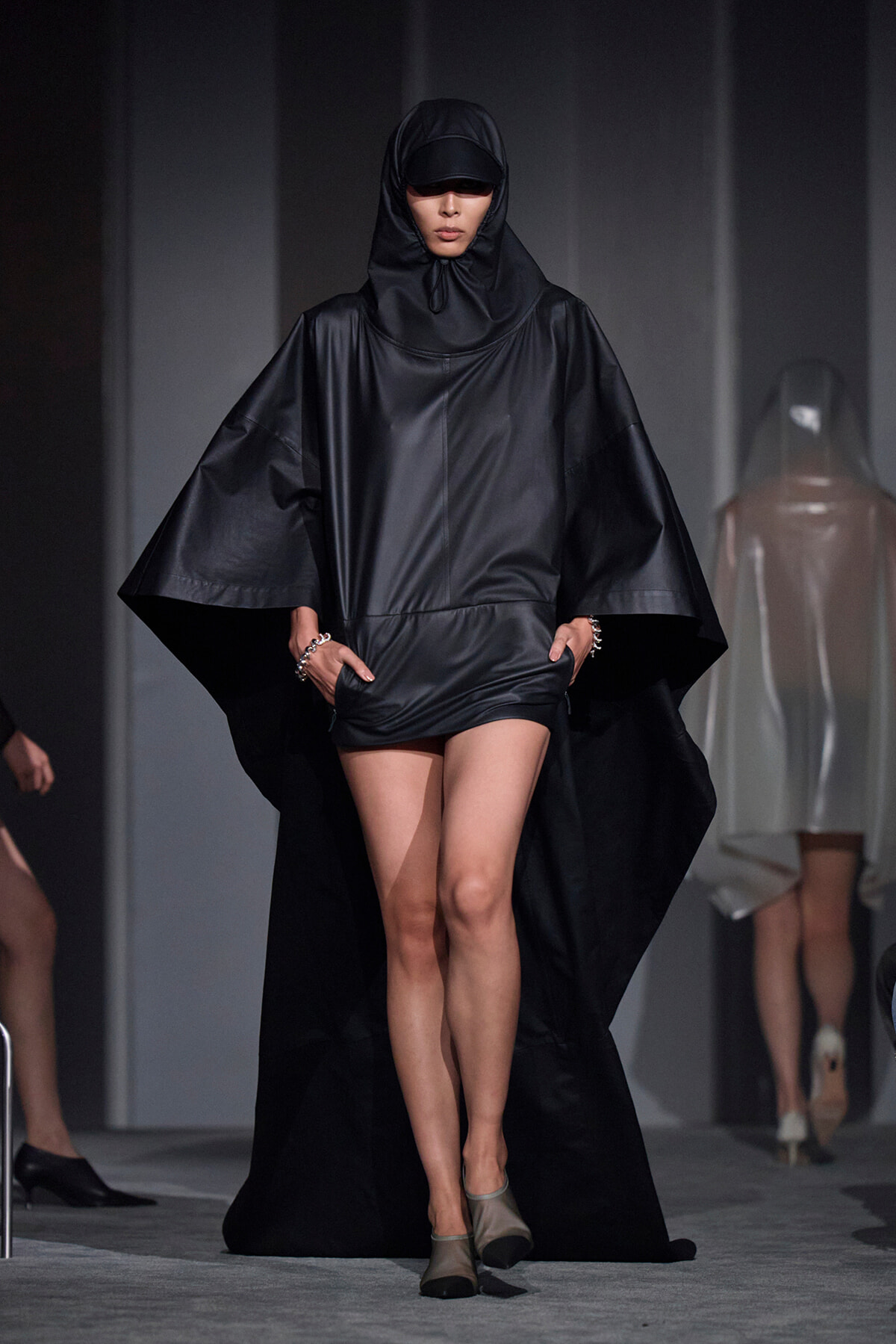 Model walking a fashion show in a black hooded cape with wide sleeves and short inner garment visible at the hembed.