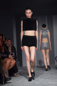 Model walks the runway in a black cropped top and matching mini skirt; another model in a silver two-piece outfit appears in the background.