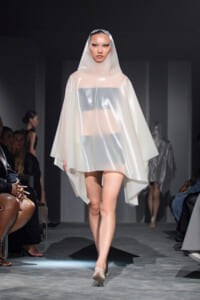 Model walks the runway in a sheer white hooded cape over a metallic bandeau top, neutral makeup, and nude heels.