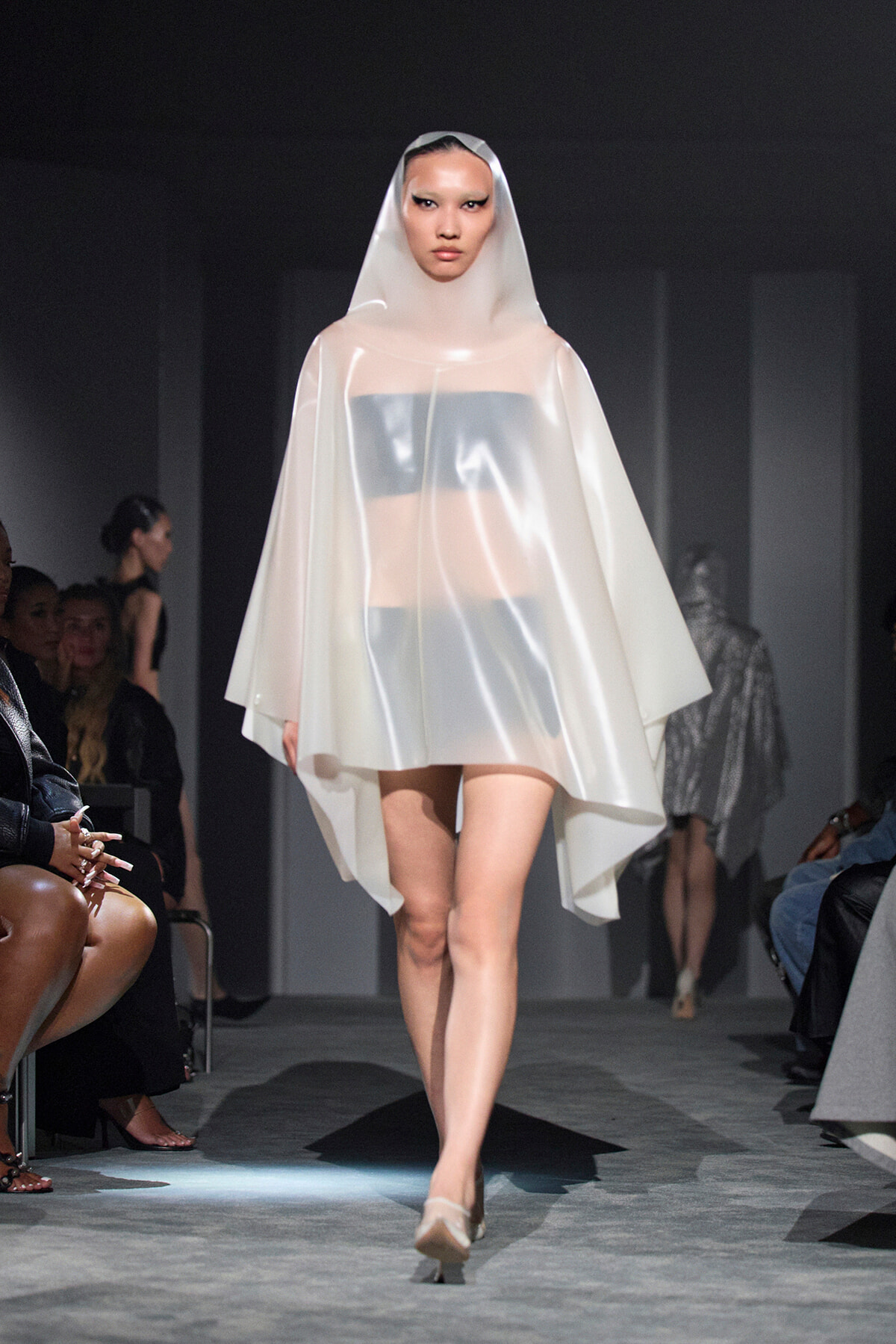 Model walks the runway in a sheer white hooded cape over a metallic bandeau top, neutral makeup, and nude heels.