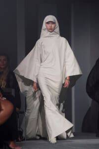 Model walking a runway in an all-white hooded cape with wide, cape-like sleeves and matching outfit.