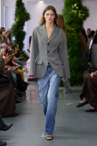 Model walks a fashion runway in a gray blazer, blue jeans, and sandals, with spectators taking photos along the sides.