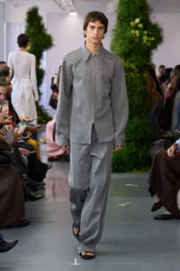 Male model in a gray zip-front shirt and wide gray pants walks the fashion show runway with spectators nearby.