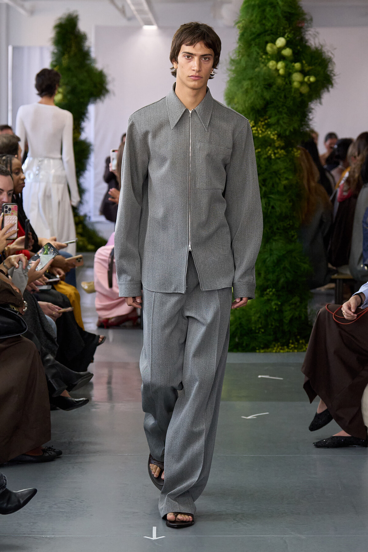 Male model in a gray zip-front shirt and wide gray pants walks the fashion show runway with spectators nearby.