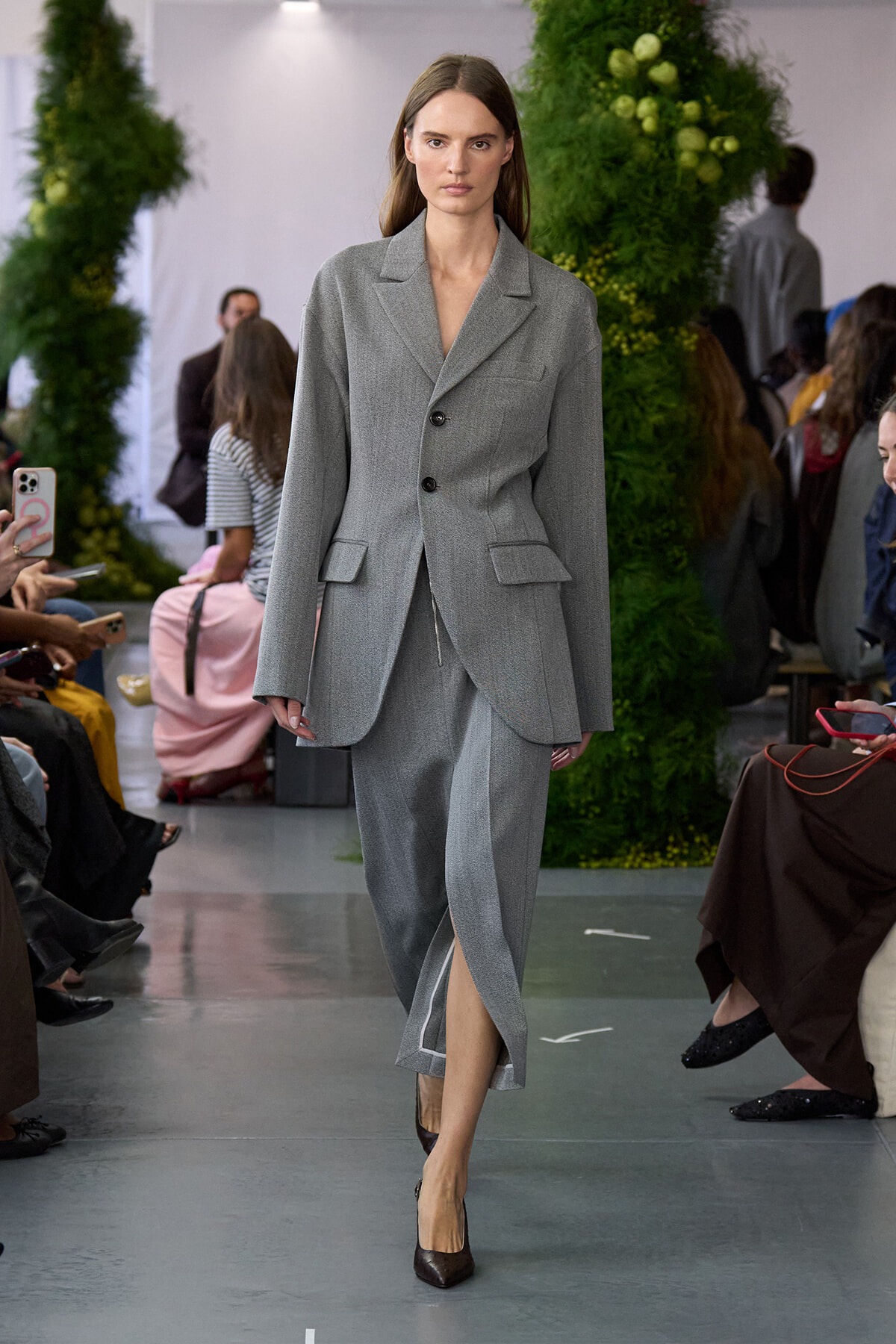 Model walks the runway in a gray tailored suit: oversized blazer with a two-button closure and matching trousers with a front slit, paired with brown heels.