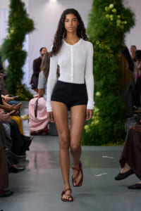 Model walking the runway in a white ribbed cardigan, black high-waisted shorts, and red sandals.