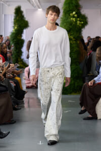 Male model walks a fashion-show runway in a white long-sleeve shirt and paint-splattered white wide-leg pants, with spectators on both sides.