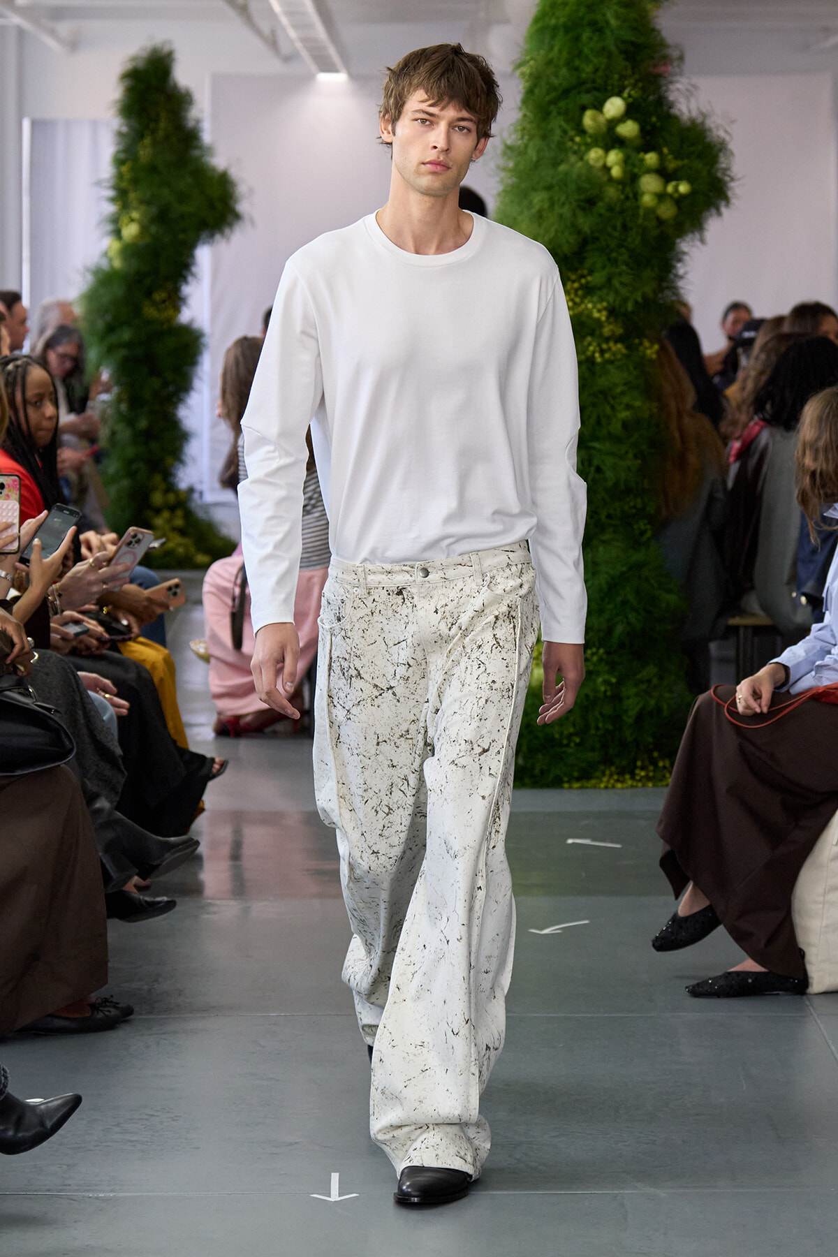 Male model walks a fashion-show runway in a white long-sleeve shirt and paint-splattered white wide-leg pants, with spectators on both sides.