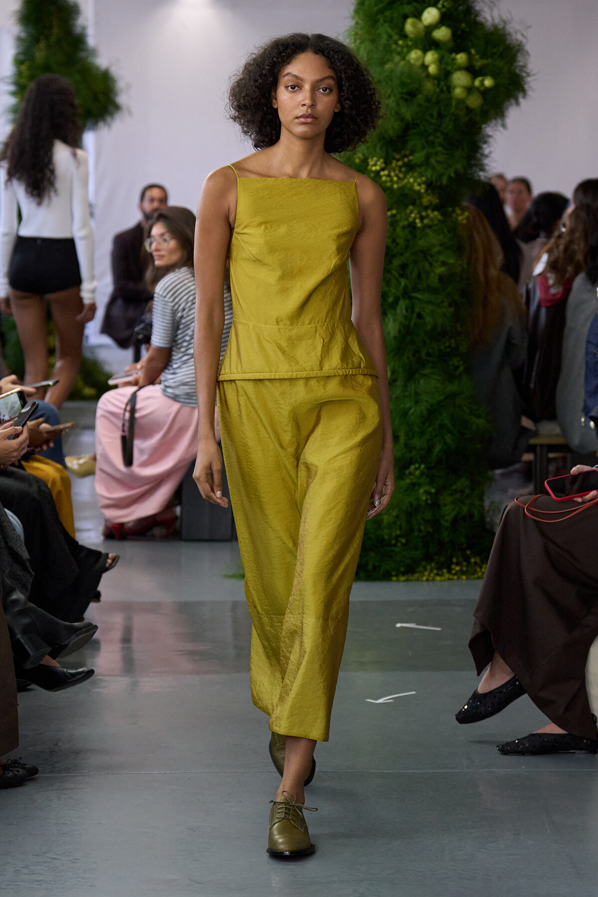 Model on a runway wearing a mustard-yellow sleeveless jumpsuit with a peplum waist and olive-green lace-up shoes, with a green plant backdrop behind.