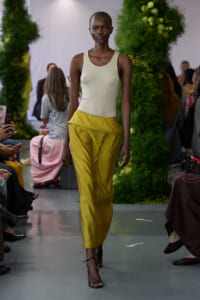 Model walks the runway in a cream sleeveless top tucked into a metallic gold skirt; green floral backdrop and seated audience on either side.