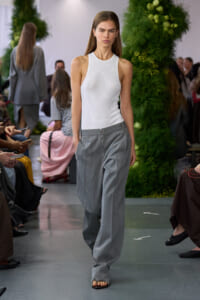 Model walking a runway in a white ribbed sleeveless top and gray wide-leg trousers, with an audience and green backdrop in the background.