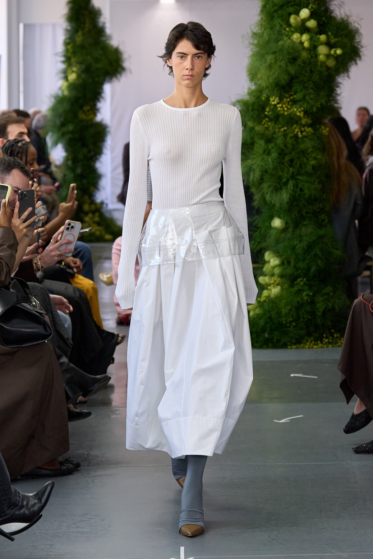 Model on a fashion runway wearing a white ribbed knit top and a voluminous white skirt with a clear plastic waist panel, surrounded by spectators and greenery.