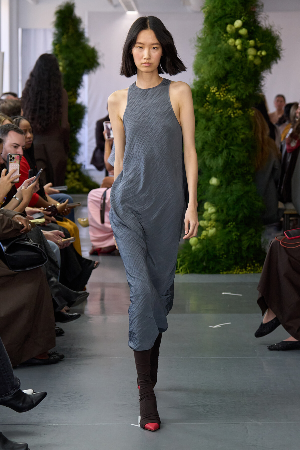 Model walks down the runway in a gray draped sleeveless dress as the audience watches and photographs the show.