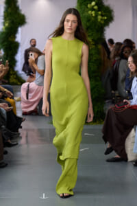 Model walks in lime-green sleeveless dress with matching wide-leg pants on a runway, audience watching.