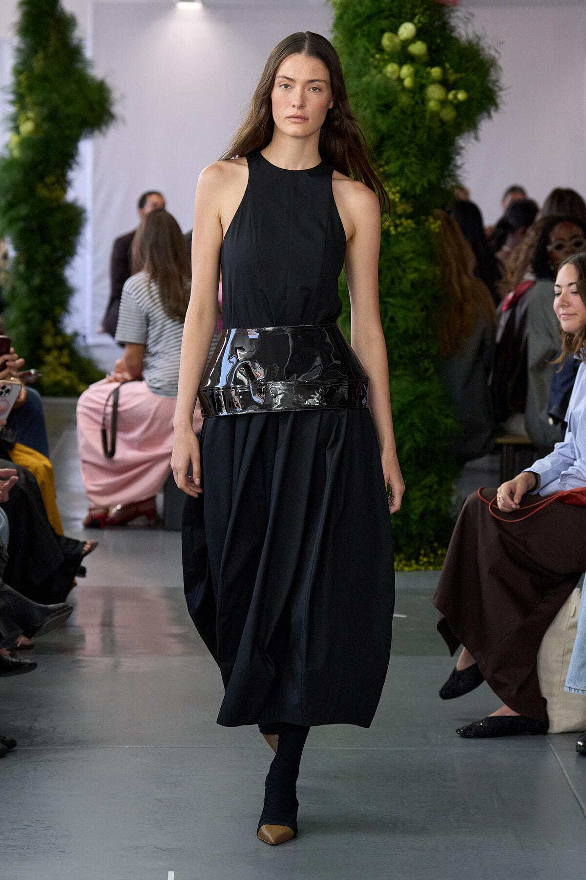 Female model walking the runway in a sleeveless black halter dress with a wide glossy belt, audience on both sides.