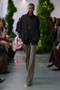 Model walking a runway in a black oversized jacket and gray wide-leg pants, audience taking photos on both sides with greenery backdrop.