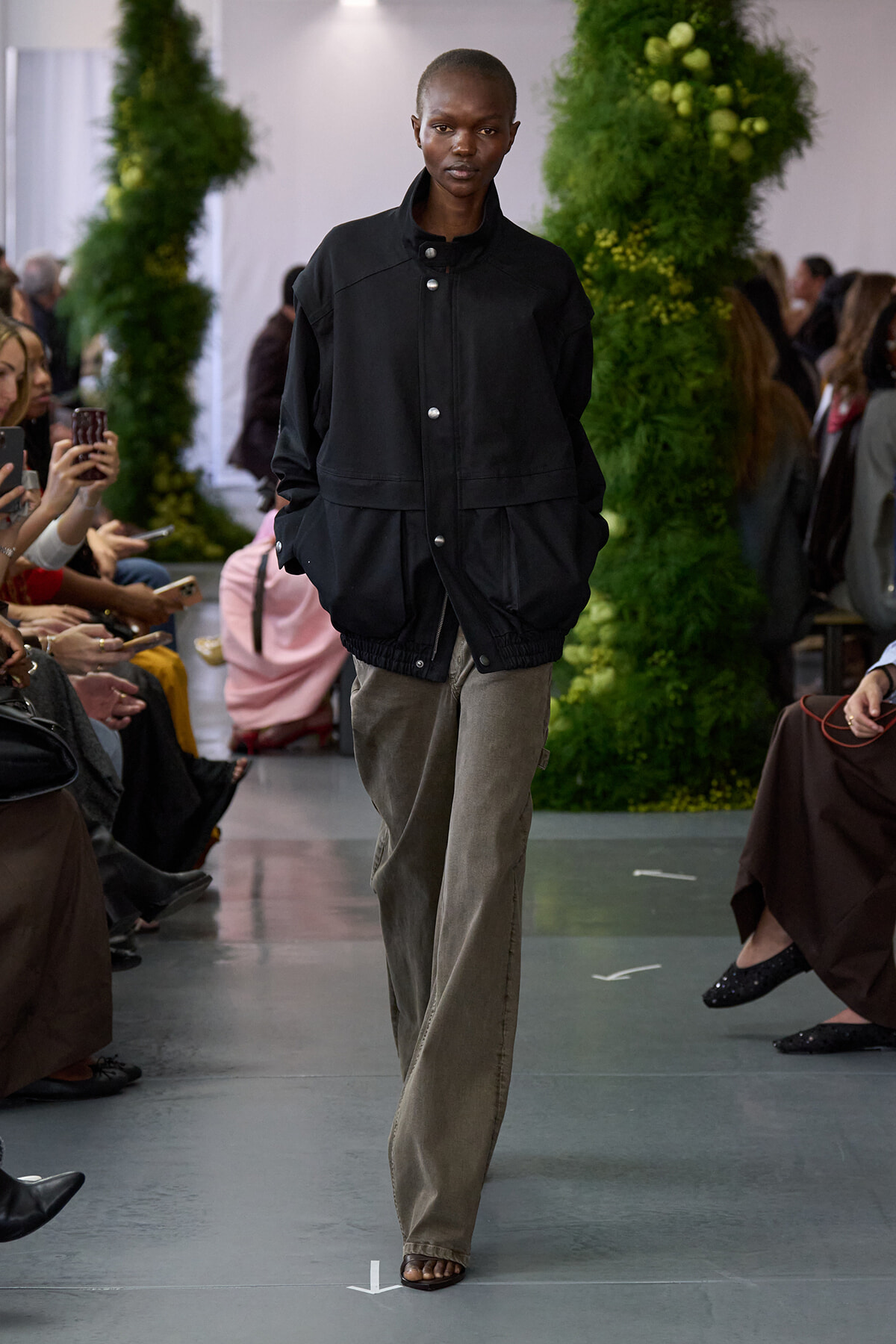 Model walking a runway in a black oversized jacket and gray wide-leg pants, audience taking photos on both sides with greenery backdrop.