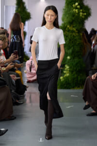 Model walking a fashion show runway in a white fitted t-shirt and black skirt with a front slit, hands in pockets.