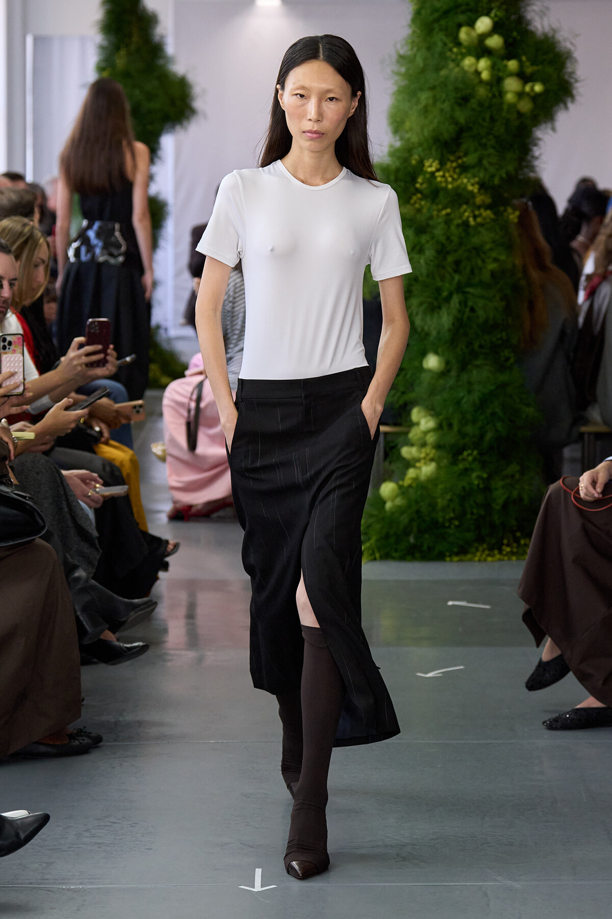 Model walking a fashion show runway in a white fitted t-shirt and black skirt with a front slit, hands in pockets.