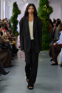 Model walking a runway in a black pinstripe suit with a cream top, audience on sides and greenery in the background