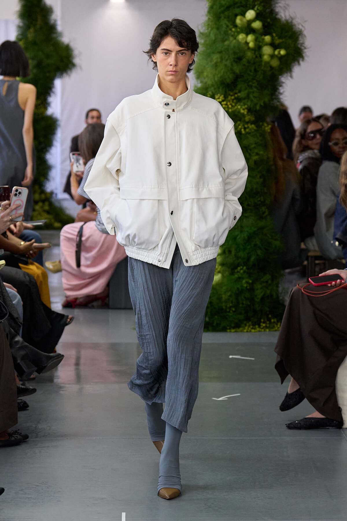 Model walking runway in a white oversized jacket and blue pleated trousers, hands in pockets, against a backdrop of green plants and an audience.