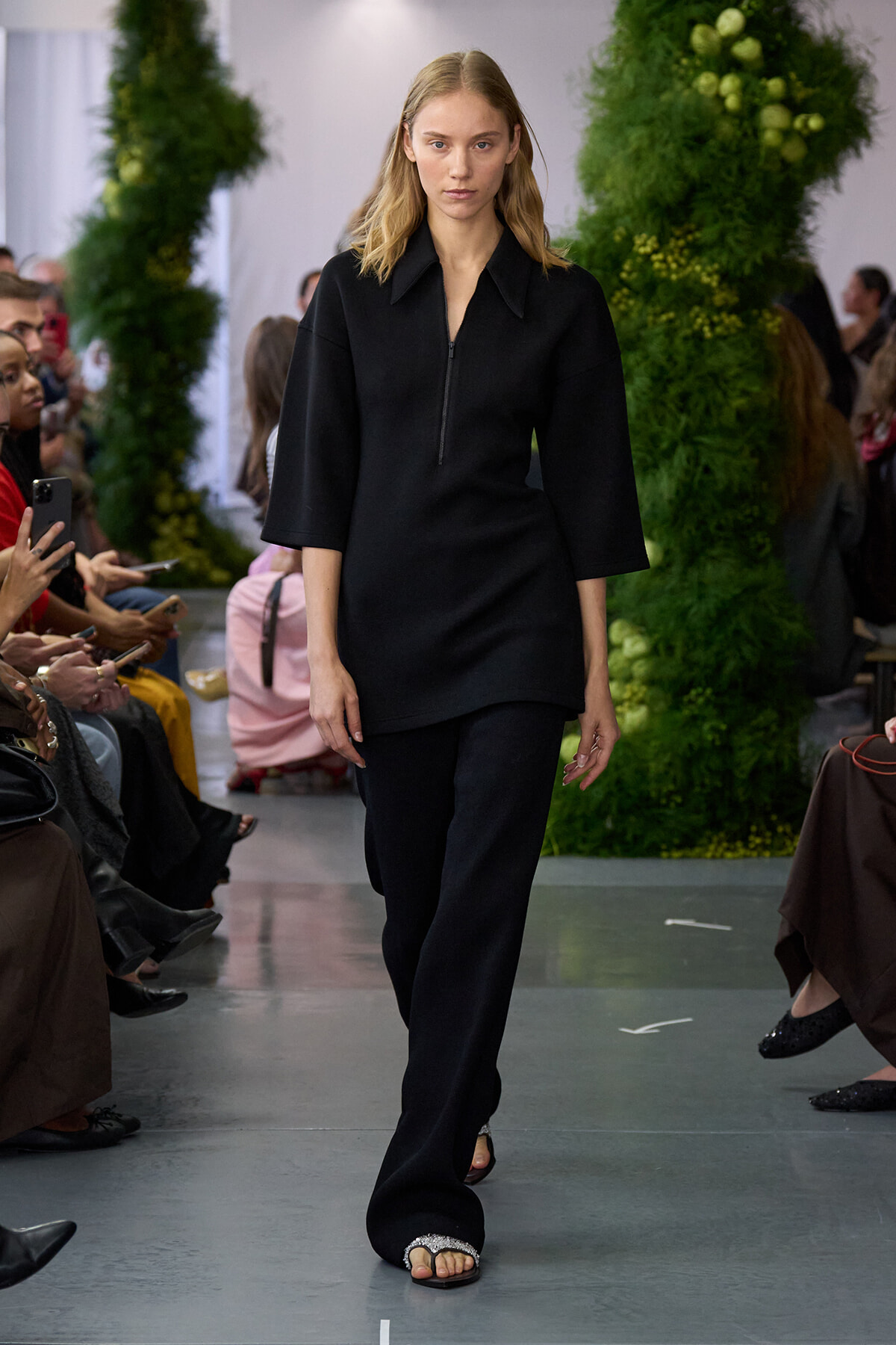 Model walks a fashion runway in a black, loose-fitting top with three-quarter sleeves and matching wide-leg pants; audience sits along the sides, greenery backdrop in the background.