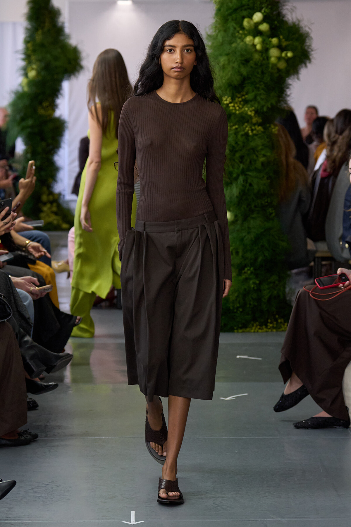 Model walking a runway in a brown ribbed long-sleeve top and dark brown pleated midi skirt, audience on both sides.