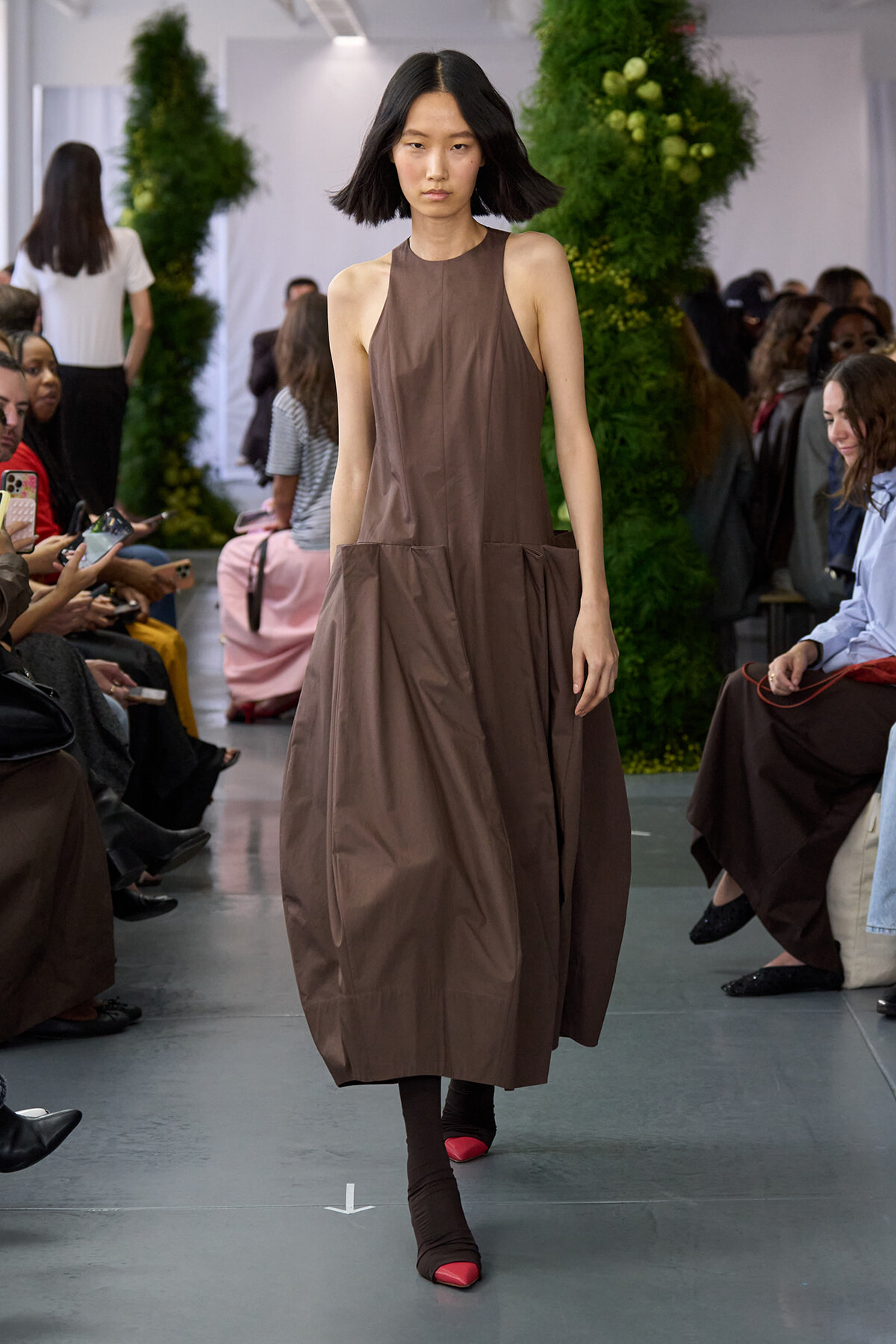 Model walking a runway in a sleeveless brown dress with oversized pockets, audience seated along the sides, greenery backdrop behind.