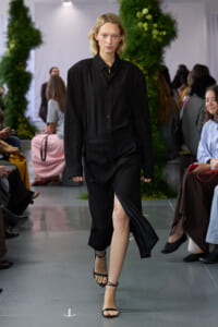 Model walks the runway in a black pinstriped shirt-dress with a front slit, wearing strappy sandals; audience seated on both sides.