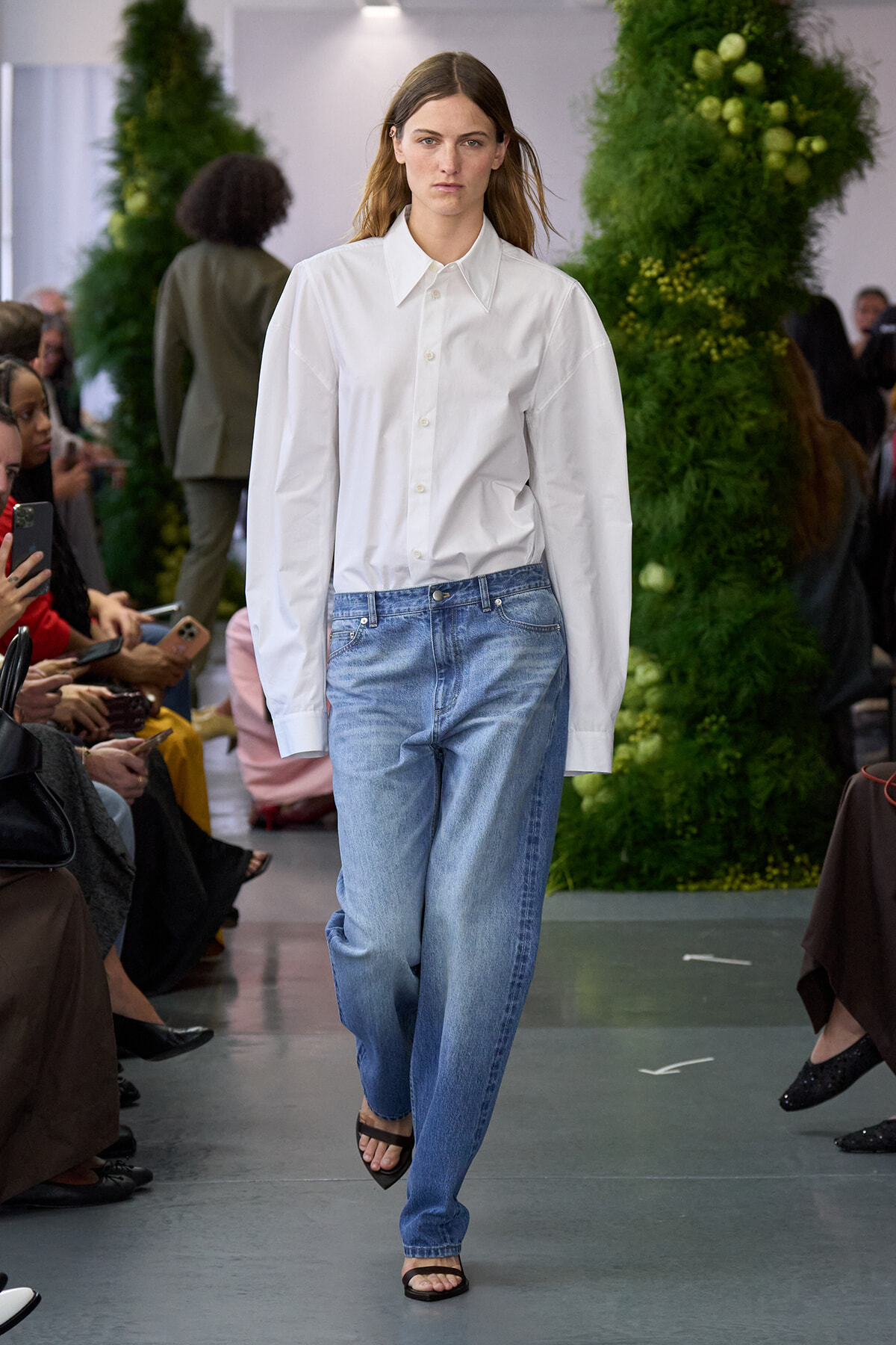 Model walking a fashion runway in an oversized white button-down shirt and light-wash jeans with black sandals.