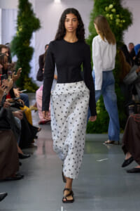 Model walking runway in a black long-sleeve top and white patterned wide-leg pants; audience seated on sides and green plants in background.