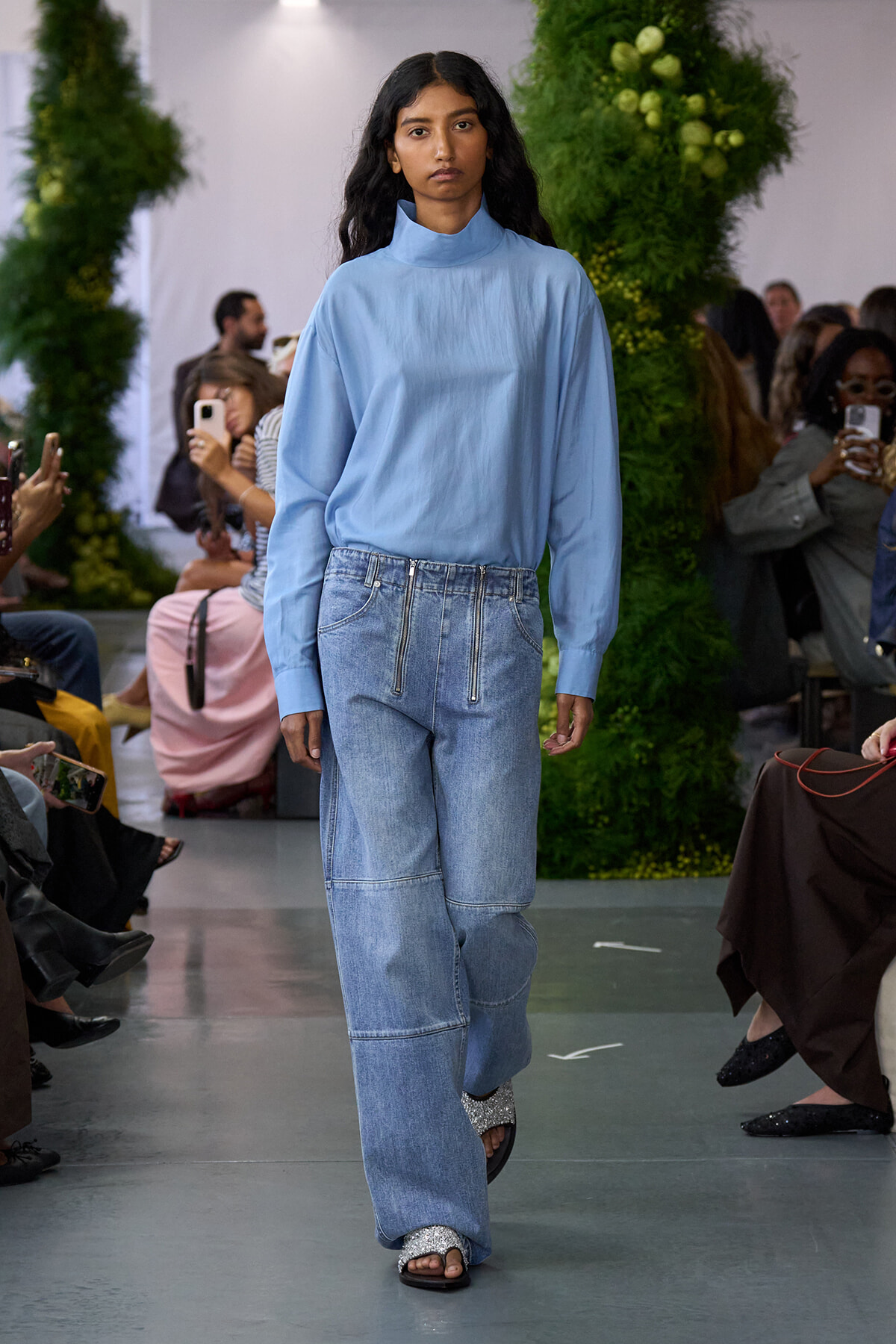 Model walks the runway in a light blue high-neck blouse and wide-leg, light-wash jeans with front zipper details.