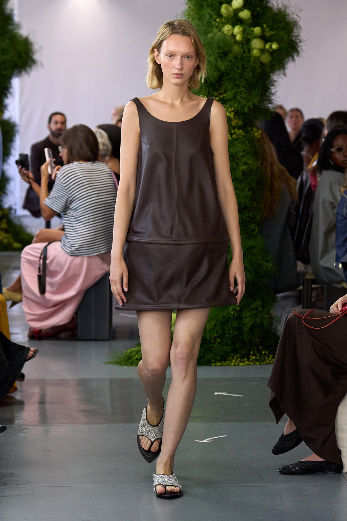 Model walking a runway in a brown sleeveless leather mini dress and glitter sandals, with green foliage backdrop.