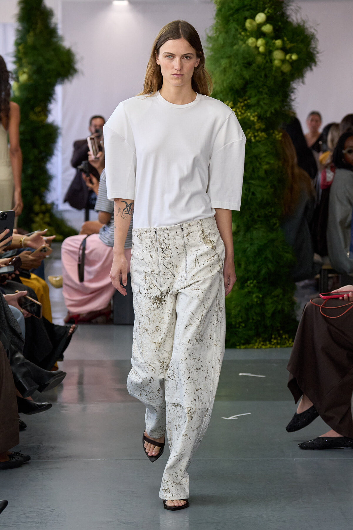 Model walks the runway in a white oversized top and splattered white wide-leg pants at a fashion show. Audience watches from sides.