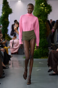 Model on a runway wearing a pink turtleneck sweater and olive high-waisted shorts, walking past a seated audience with green decor behind.