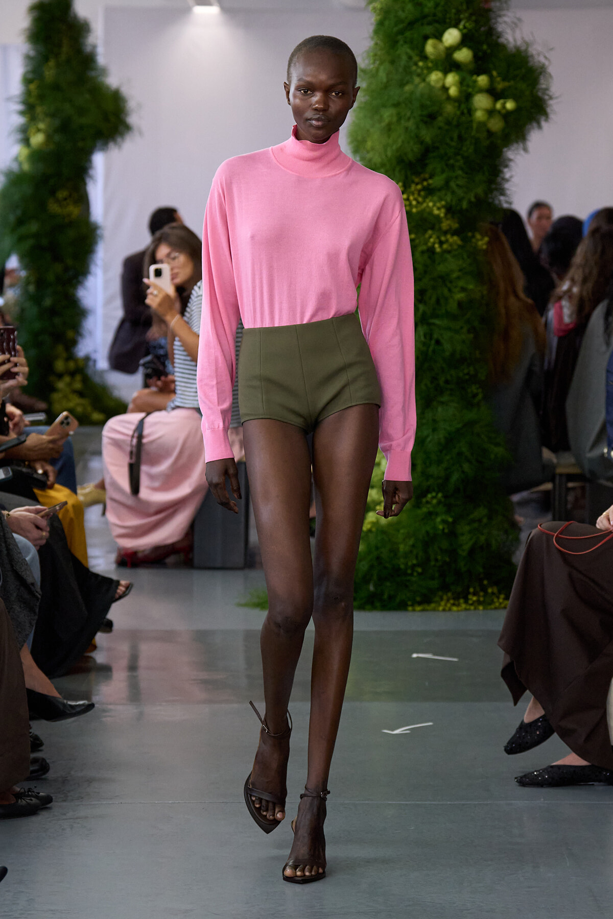 Model on a runway wearing a pink turtleneck sweater and olive high-waisted shorts, walking past a seated audience with green decor behind.
