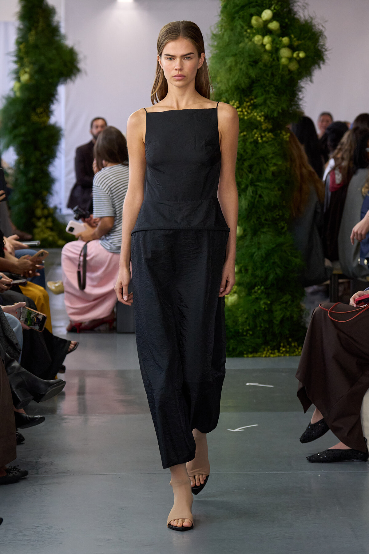 Model on a runway wearing a sleeveless black midi dress with thin straps, beige open-toe sandals, and seated audience on both sides.