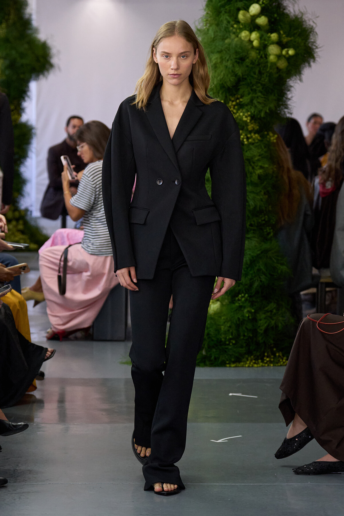 Model walking the runway in a black double-breasted tailored suit with wide-leg pants and open-toe sandals; green plant backdrop and audience visible.