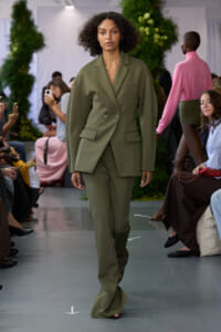 Model walking runway in an oversized olive-green suit with double-breasted blazer and wide-leg pants, audience on both sides.