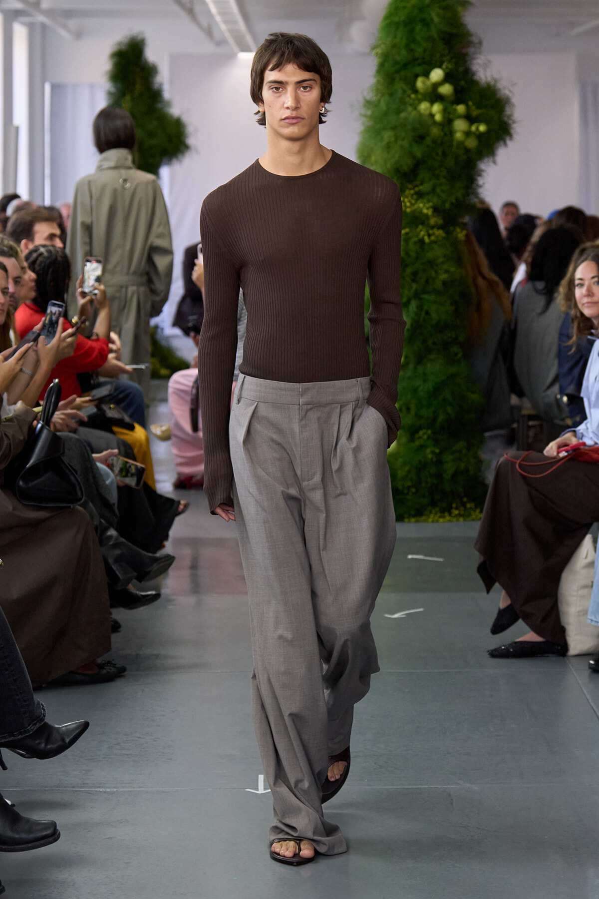 Male model walks the runway in a brown knit top and loose gray trousers, flanked by seated audience.