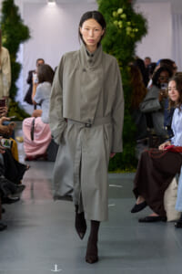 Model walks the runway in a light gray belted trench coat with a high collar.