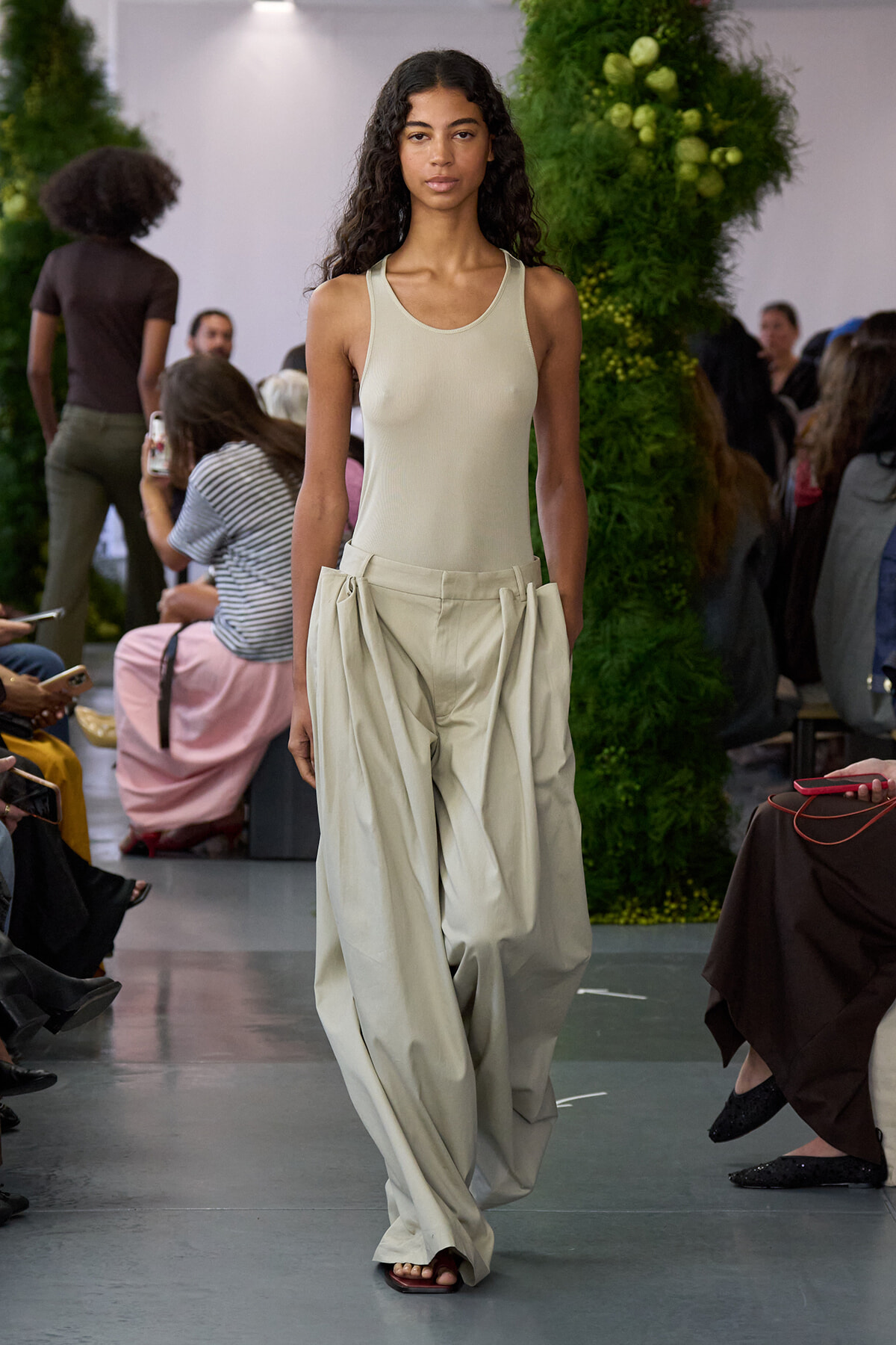 Model walks the runway in a cream sleeveless top and oversized beige pleated pants, with audience in the background.