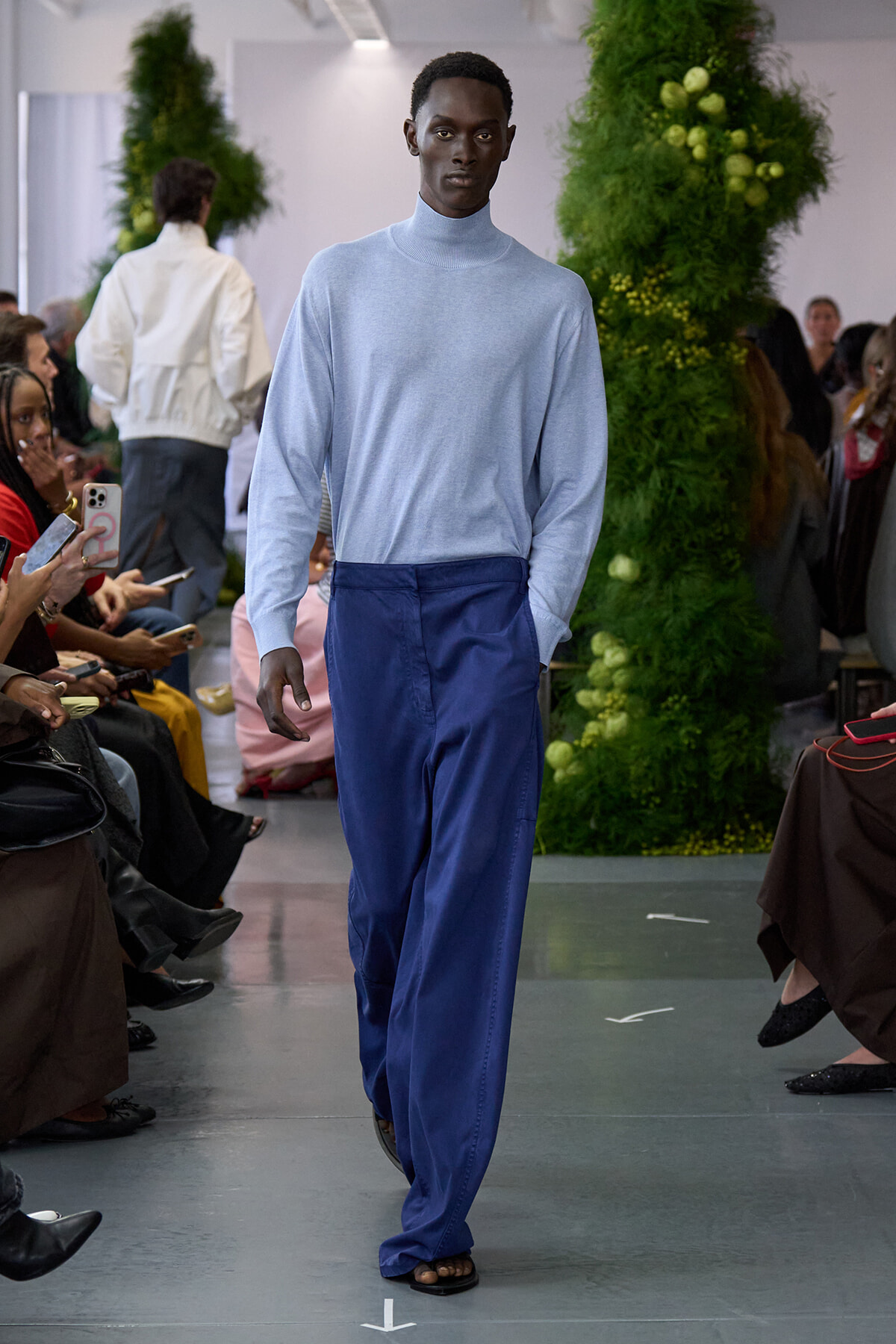 Male model walks a fashion runway wearing a light blue turtleneck sweater and royal blue wide-leg pants, audience on both sides watching the show.