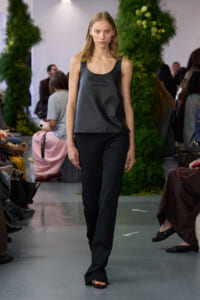 Model walking a fashion runway in a dark gray sleeveless top and black trousers, audience on both sides, green decor in background.