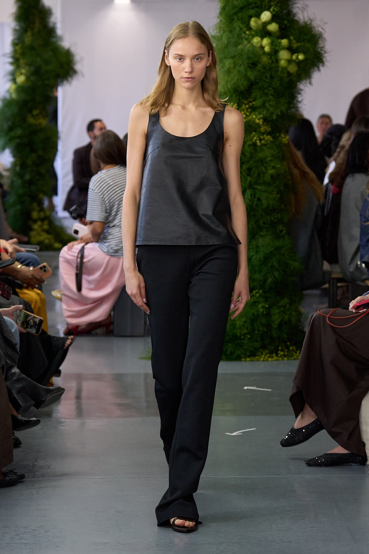 Model walking a fashion runway in a dark gray sleeveless top and black trousers, audience on both sides, green decor in background.