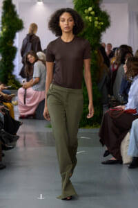 Model walking a runway in a brown fitted top and olive green wide-leg pants, audience seated on both sides against greenery backdrop.