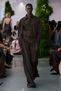 Male model walking the runway in a dark brown button-up shirt and wide-leg brown trousers at a fashion show