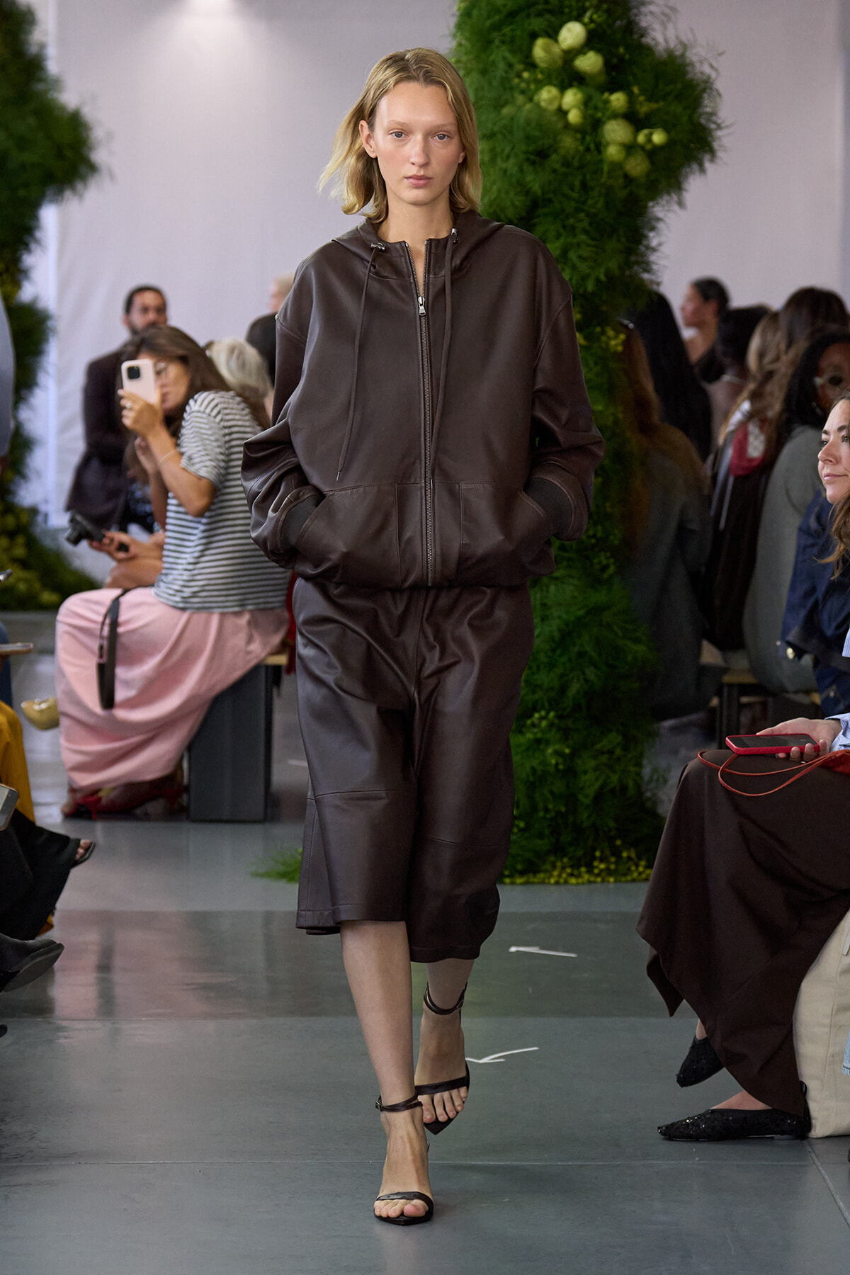 Model walks the runway in a dark brown leather hooded zip-up jacket with matching knee-length shorts and strappy sandals.