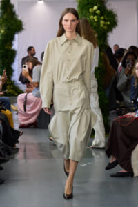 Model on a runway wearing a beige button-down shirt with high-waisted, sculpted cropped pants and brown heels.